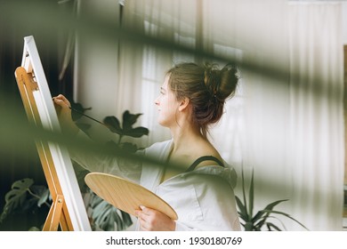 Side View Of Attractive Woman In White Shirt Using Brush And Wooden Palette While Painting On Canvas In Studio. Art Therapy Concept.