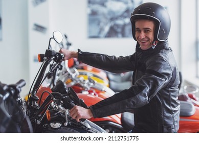 Side View Of Attractive Young Blond Man In Black Leather Jacket And Helmet Looking At Camera And Smiling While Sitting On A Motorbike