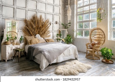 Side view of authentic cozy bedroom with interior design in boho chic style, decorative headboard over comfort bed, wicker armchair, houseplants near commode and mirror. Lovely room in classic house