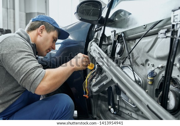 Side view of auto\
mechanic in blue uniform and cap repairing electrical wires in door\
of old car. Middle aged man changing damaged details to new ones in\
car service station