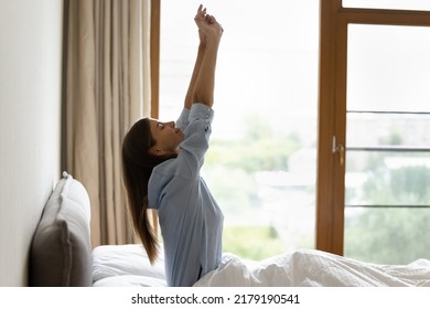 Side view awakened woman sit on bed, rise arms up, stretch back feels refreshed after enough healthy night sleep on orthopaedic mattress on window background. Good morning, welcoming new day concept - Shutterstock ID 2179190541