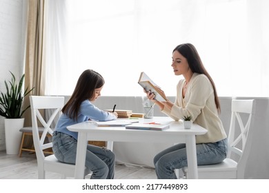 Side View Of Babysitter Reading Aloud Near Girl Writing Dictation At Home