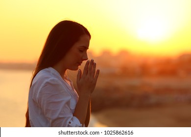 Side view backlight portrait of a single woman praying and looking down at sunset