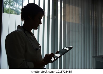 Side View Backlit Silhouette Of Female Doctor In Medical Coat Standing Against Window In Healthcare Office With Stethoscope Around Her Neck And Using Tablet Computer