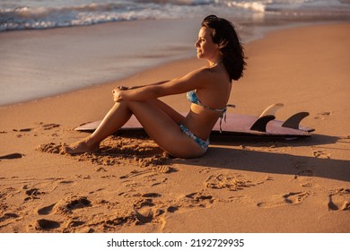 Side View Of Barefoot Hispanic Woman In Bikini Sitting On Sand Near Surfboard And Admiring Sea While Spending Summer Weekend Day On Beach