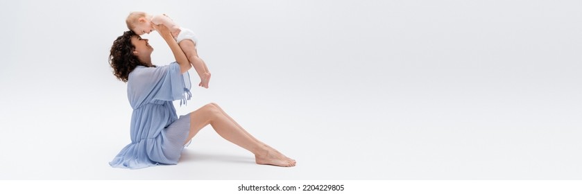 Side View Of Barefoot Parent In Dress Holding Cheerful Infant Kid On White Background, Banner