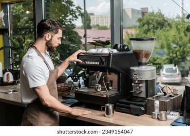 side view of bearded barista in apron making coffee on coffee machine near pitchers in cafe - Powered by Shutterstock