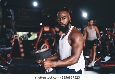 Side view of bearded black male athlete in sportswear sitting with heavy dumbbells on knees while looking at camera during training in gym over blurred background of people and sports facilities - Powered by Shutterstock