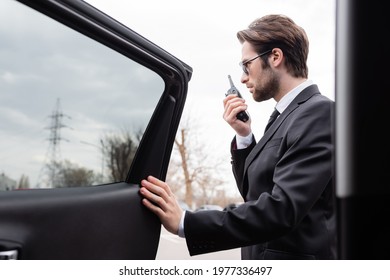 side view of bearded bodyguard in suit and sunglasses using walkie talkie near modern car - Powered by Shutterstock