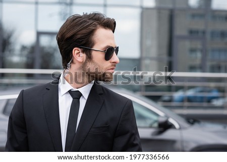 side view of bearded bodyguard in suit and sunglasses with security earpiece near modern car