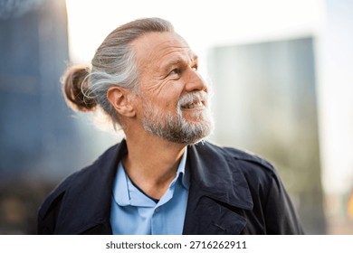 Side view of a bearded elderly man with gray hair in a bun smiling thoughtfully while looking into the distance, dressed casually outdoors