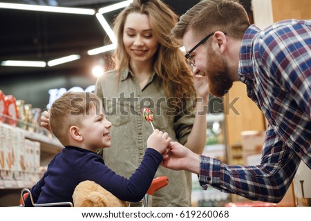 Side view of a bearded father in eyeglasses giving a candy to his son which sitting in shopping trolley in supermarket while mother standing near them