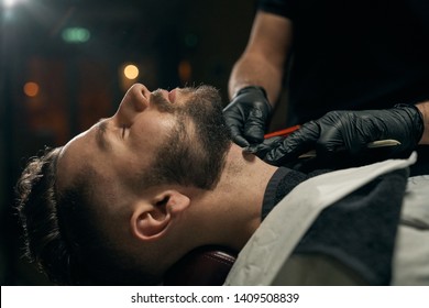 Side view of bearded handsome guy lying on couch while competent male barber in black gloves shaving his beard by sharp razor.Young client at barbershop is changing hairstyle and appearance