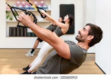 Side View Of Bearded Male Athlete Smiling And Doing Low Row Exercise With TRX Ropes During Group Suspension Training In Gym