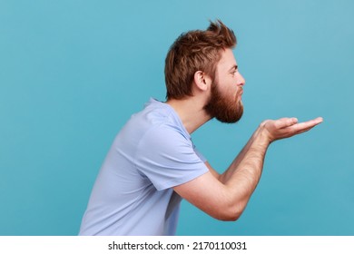 Side view of bearded man blowing air kiss keeps lips, folded palm towards mouth, sends mwah coquettish, wants to cheer up her partner. Indoor studio shot isolated on blue background.