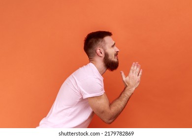 Side view of bearded man folding hands in prayer, closing eyes and talking to god, asking for help, expressing gratitude, wearing pink T-shirt. Indoor studio shot isolated on orange background.