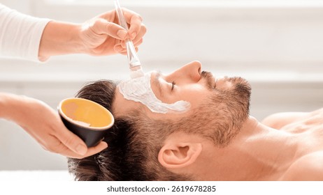 Side view of bearded man getting face treatment at modern spa salon. Female attendant applying nourishing face mask on sleeping middle aged man face. Male cosmetology and face care concept