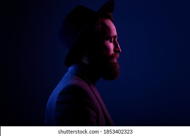 Side view of a bearded man in hat and suit, posing in the studio, studio lights, isolated on dark blue background.