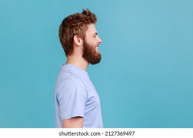 Side View Of Bearded Man Looking At Camera With Toothy Smile And Happy Facial Expression, Being In Good Mood, Rejoicing Great Results. Indoor Studio Shot Isolated On Blue Background.