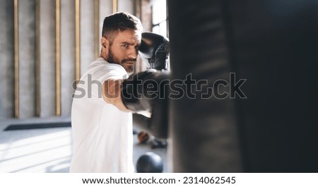 Side view of bearded man looking at camera and hitting punching bag with boxing gloves while training in sunlit gym