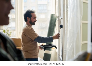 Side view of bearded man with paintroller applying paint on wall during renovation of domestic room of new apartment