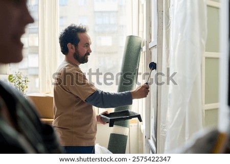 Side view of bearded man with paintroller applying paint on wall during renovation of domestic room of new apartment