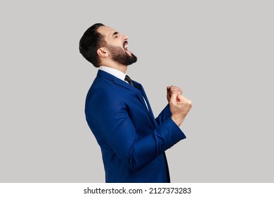 Side view of bearded man showing yes gesture and screaming celebrating his victory, success, dreams comes true, euphoria, wearing official style suit. Indoor studio shot isolated on gray background.