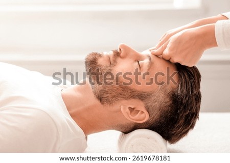Side view of bearded middle aged man having relaxing head massage at white modern spa. Unrecognizable spa attendant rubbing sleeping man head, making healing head massage, male spa concept