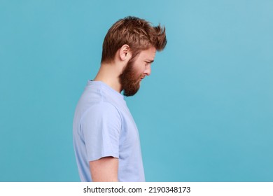 Side view of bearded upset young man standing and looking at camera with dissatisfied sadness face, expressing sorrow, having bad mood. Indoor studio shot isolated on blue background.