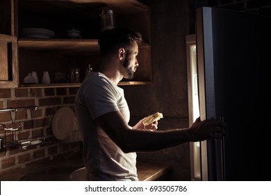 Side View Of Bearded Young Man In Pajamas Eating And Looking At Refrigerator At Night