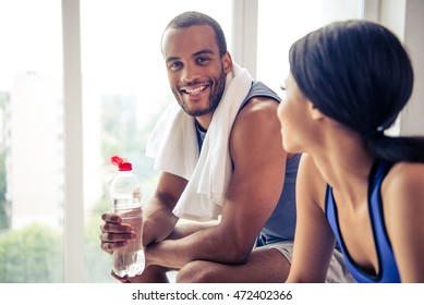 Side View Of Beautiful Afro American Couple In Sports Clothes Resting After Workout. Guy Is Holding A Bottle Of Water, Looking At Camera And Smiling