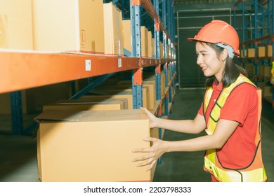 Side View Beautiful Asian Female Worker Working The Night Shift Wearing Helmet Doing Her Job With Smiling Face, Checks And Lifts Boxes From Shelves In A Warehouse To Keep Things Tidy.