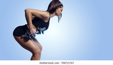 Side view, beautiful athletic young woman, bodybuilder lifting weights, blue gradient background ; copy space 