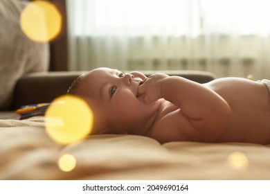 Side view of beautiful baby boy without clothes, lying on back, exploring its mouth by fingers and smiling. Teething baby, selective focus with bokeh circles.