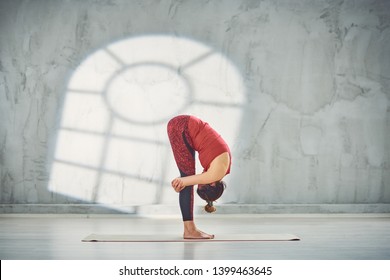 Side view of beautiful Caucasian brunette in red sports wear standing barefoot on the mat in Gorilla yoga pose.