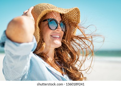 Side view of beautiful mature woman wearing sunglasses at beach. Young smiling woman on vacation looking away while enjoying sea breeze wearing straw hat. Closeup portrait of attractive girl relaxing.