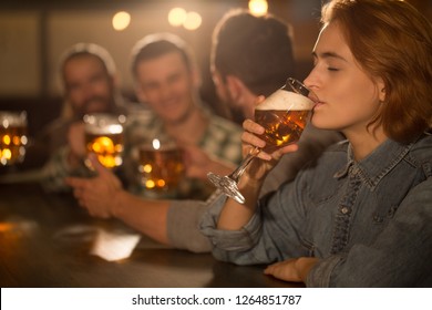 Side view of beautiful woman holding glass of tasty beer and drinking. Young woman with closed eyes enjoying and tasting beer in bar. People resting and communicating on background.