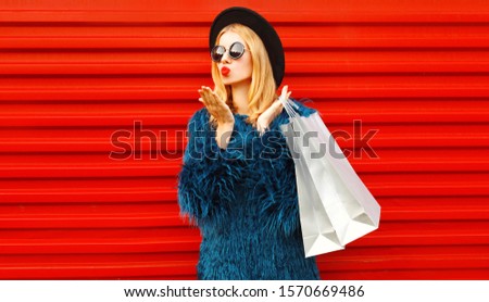 Side view beautiful woman with shopping bags blowing red lips sending sweet air kiss, stylish female model wearing blue faux fur coat, round hat and sunglasses over red wall background