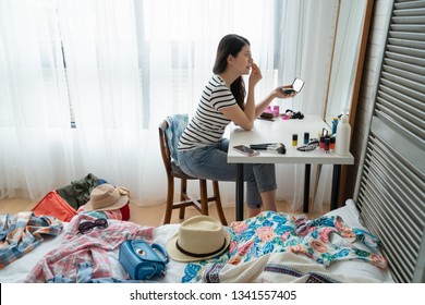 Side View Beautiful Young Asian Woman Sitting At Dressing Table And Doing Her Makeup Looking In Mirror. Messy Bed Room While Packing Luggage For Spring Vacation Holidays To Beach With Hat And Cloth.