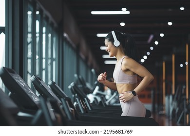 Side view of beautiful young asian woman running on treadmill and listening to music via headphone during sports training in a gym. - Powered by Shutterstock