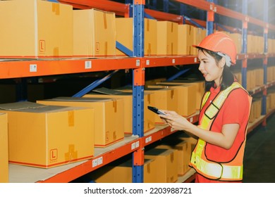 Side View Beautiful Young Asian Female Worker Working The Night Shift Wearing A Helmet Works With Smiling Face, Use Tablet To Inspect The Boxes From The Warehouse Shelf To Verify They Are Correct.