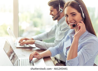 Side view of beautiful young business woman and handsome businessman in headsets using laptops while working in office. Girl is looking at camera