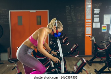 Side view of beautiful young Caucasian female athlete with oxygen mask exercising with exercise bike in fitness studio. Bright modern gym with fit healthy people working out and training - Powered by Shutterstock