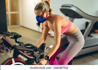 Side view of beautiful young Caucasian female athlete with oxygen mask exercising with exercise bike in fitness studio. Bright modern gym with fit healthy people working out and training - Powered by Shutterstock