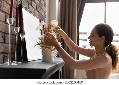 Side view beautiful young latin woman decorating fireplace with dried flowers in vase, enjoying improving or styling apartment interior, arranging indoors decor in living room, coziness concept.