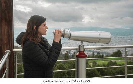 Side view of beautiful young woman, standing at a viewing platform, holding in her hands long binoculars mounted on a metal pedestal against a cloudy sky background - Powered by Shutterstock