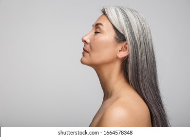 Side View Beauty Portrait Of An Attractive Sensual Mature Topless Woman With Long Gray Hair Standing Isolated Over Gray Background, Eyes Closed