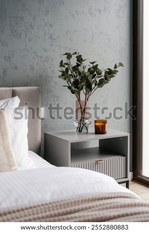 Side view of bedside table with candle and home decor next to neatly made bed in hotel rom. Comfort bedroom interior with eucalyptus plants in vase on nightstand. Quiet living, styling spaces concepts