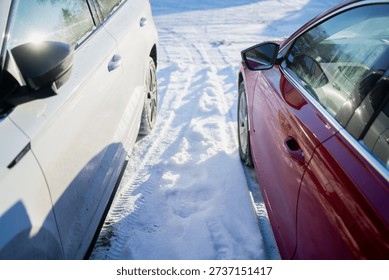 Side view between white and red cars parked on snowy winter driveway, tire tracks and packed fresh snow in bright sunlight, close up of vehicle doors and side mirrors outdoors
