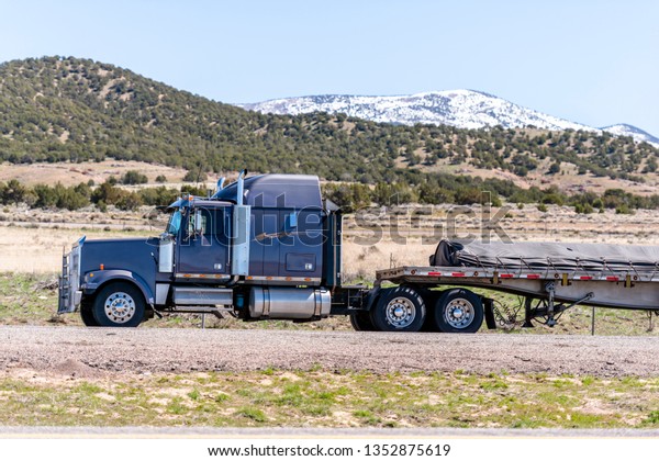 Side\
view of big rig classic bonnet American dark blue long haul semi\
truck transporting covered cargo on flat bed semi trailer going on\
divided highway in Utah with mountain on\
background